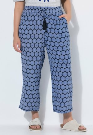 Person, die blaue gemusterte Hosen mit weitem Bein und schwarzen Kordelquasten sowie beige Schlupfsandalen trägt, stehend vor weißem Hintergrund.