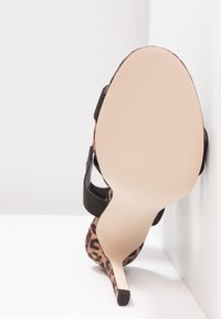 Sandale à talons hauts imprimé léopard avec semelle beige et brides noires, photographiée par le dessous sur un fond blanc d'angle.