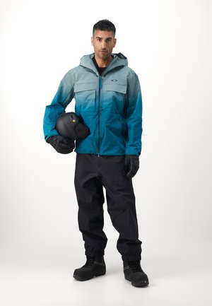 Férfi két színű kék vízálló dzseki kapucnival, fekete nadrág, kesztyű és fekete sisak, amely a funkcionális dizájnt és az anyag textúráját hangsúlyozza.