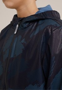 Černá síťovaná bunda s modrými akcenty a kapucí. Obsahuje přední zip a lehkou, prodyšnou texturu. Vzor zobrazuje abstraktní tvary.