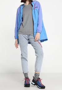 Blauer und lila Windbreaker mit Kapuze, grauer Sweatshirt, helle Jeans und schwarze Wanderstiefel mit blauen Akzenten und pinken Schnürsenkeln.
