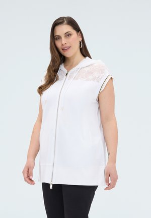 Kobieta o długich brązowych włosach nosi białą bezrękawnikową bluzę z kapturem z kwiatową koronkową wstawką i czarne spodnie, stojąc na jednolitym tle.