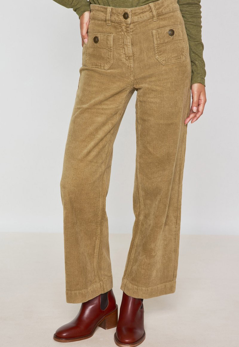Pantaloni a gamba larga in velluto a coste di colore marrone chiaro con tasche frontali e dettagli con bottoni, abbinati a stivaletti con tacco marroni.