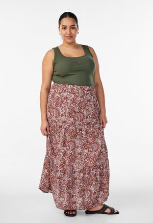 Žena stoji, nosi maslinasto zeleni top, dugu cvjetnu paisley suknju u toplim tonovima i crne sandale na bijeloj pozadini.