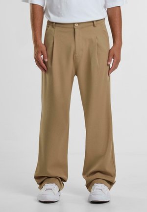 Man draagt loszittende, kaki-kleurige broek met plooien, gecombineerd met witte sneakers en een in de broek gestoken wit overhemd.
