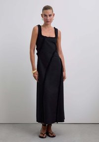 Zwarte maxijurk met een vierkante halslijn, getextureerde ruches en een vloeiende silhouet. Gepaard met zwarte sandalen.