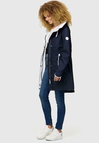 Marinblå vattentät jacka med en vitrandig huva, framsida med dragkedja, fickor och en lös passform, ihop med blå jeans och vita sneakers.
