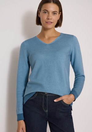 Femme portant un pull bleu clair à manches longues avec col en V et un jean bleu foncé, la main droite dans la poche, debout devant un mur uni.