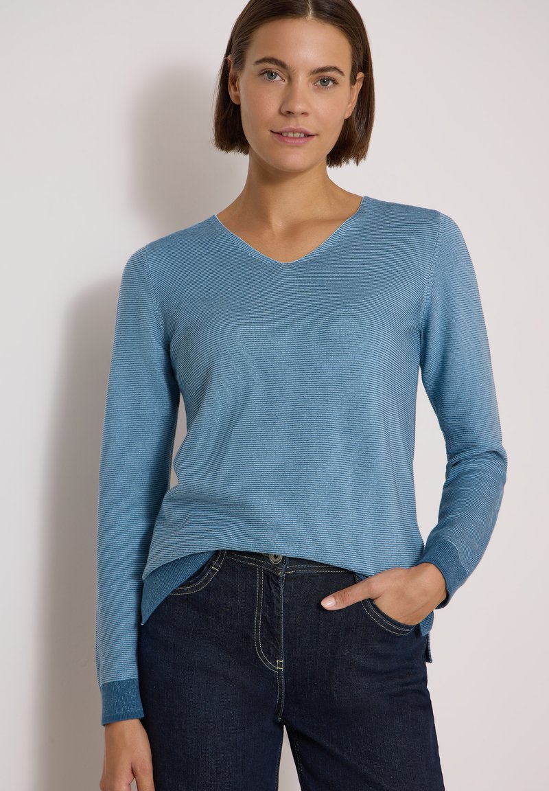 Femme portant un pull bleu clair à manches longues avec col en V et un jean bleu foncé, la main droite dans la poche, debout devant un mur uni.