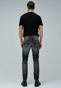 Camisa negra de manga corta combinada con jeans ajustados de color gris oscuro que presentan una etiqueta azul, bolsillos traseros y patrones claros deslavados. Zapatos negros.