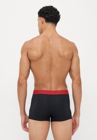 Schwarze Boxershorts mit einem roten elastischen Bund, die eine glatte Textur und einen engen Sitz bieten. Von hinten betrachtet.