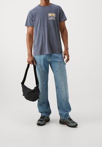 Blue crew-neck t-shirt with graphic print, light blue jeans with side pockets, and black crossbody bag. Gray-and-black sneakers with accents.
