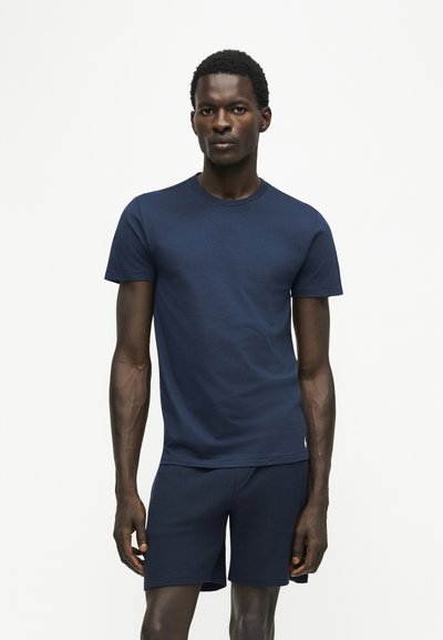 Homme aux cheveux courts et bouclés portant un t-shirt bleu marine uni à manches courtes et un short bleu marine assorti, debout devant un fond blanc.
