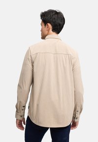 Camisa de pana beige con una textura suave, puños con botones y un diseño liso en la parte de atrás. Presenta un cuello clásico y un corte relajado.