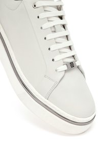 Sneaker bianca in pelle con suola spessa, lacci bianchi e un piccolo dettaglio metallico vicino all'occhiello inferiore su uno sfondo semplice.
