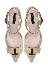 Zapatos de tacón en tono beige con punta afilada, que presentan una textura trenzada, detalles en dorado y correas ajustables en el tobillo. El logo es visible en la plantilla.
