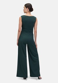 Zöld overál, amely illeszkedő felsőrésszel, széles lábakkal, sima anyaggal és rejtett hátsó cipzárral rendelkezik. Minimalista dizájn, letisztult vonalakkal.