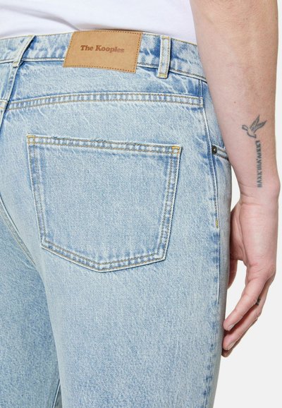 Jean en denim bleu clair avec une poche arrière et une étiquette en cuir marron "The Kooples", montrant un avant-bras et une main tatoués reposant à côté.
