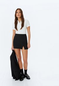 Camisa polo blanca de manga corta, falda mini negra con abertura lateral, blazer negro colgado del brazo, calcetas negras cortas, zapatos brillantes con acentos dorados.