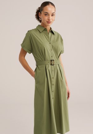Frau trägt ein olivgrünes, kurzärmeliges Midikleid mit Knopfleiste und passendem Gürtel, die Hände in den Taschen, vor einfarbigem Hintergrund.