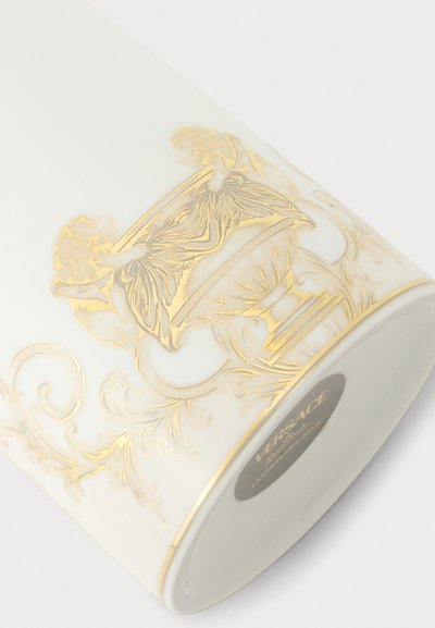 Tasse en céramique blanche avec un motif ornemental doré complexe et une étiquette Versace au fond.