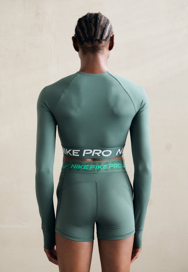 Top atletico verde con maniche lunghe e pantaloni corti aderenti, con una vita a contrasto nera e verde con marchio. Tessuto liscio ed elastico.