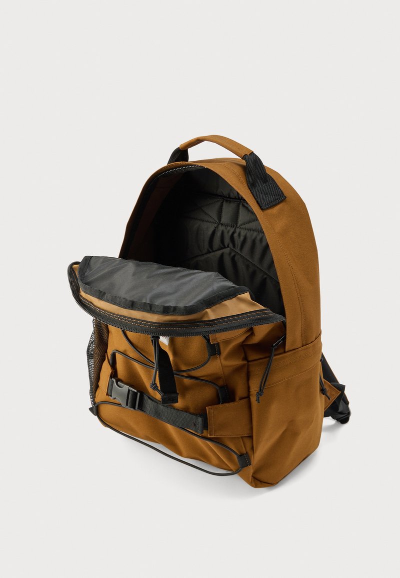 Sac à dos en tissu marron avec des accents noirs, présentant un compartiment principal zippé, un rabat supérieur ouvert et des bretelles réglables.