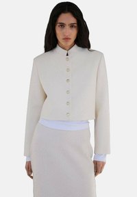 Veste crème courte avec un col haut, ornée de sept boutons dorés, assortie à une jupe texturée de couleur claire et à des manches longues blanches.