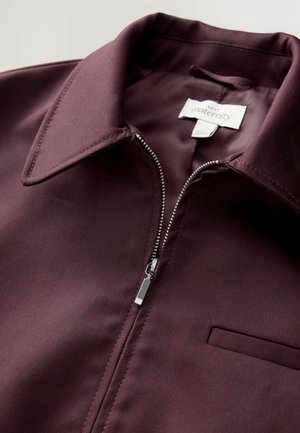 Bordo jakna za nosečnice z zadrgo in zložljivim ovratnikom, gladka tekstura, srebrna zadrga in majhna žepna obloga na levi strani.