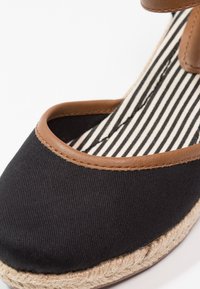 Gros plan d'une chaussure espadrille noire avec des bordures brunes, un intérieur rayé noir et blanc, et une semelle en jute tressé sur fond blanc.