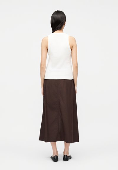 Femme aux longs cheveux bruns portant un haut blanc sans manches, une jupe midi marron et des chaussures plates noires, debout de dos sur un fond uni.