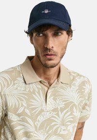 Marinekasket med broderet logo, parret med en beige polo med hvide tropiske blade og ananas mønstre. Blødt stof med krave design.