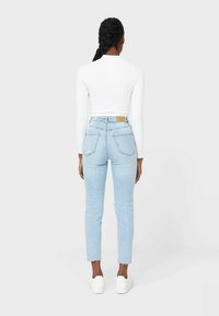 Mujer con el cabello trenzado, vistiendo una blusa blanca de manga larga, jeans de tiro alto de color azul claro y zapatillas blancas, de pie de espaldas.