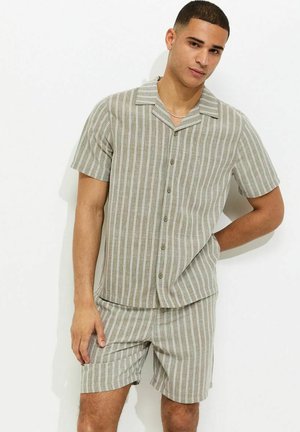 Młody mężczyzna ubrany w pasującą beżowo-białą koszulę w pionowe paski na krótki rękaw oraz krótkie spodenki, stoi na białym tle.