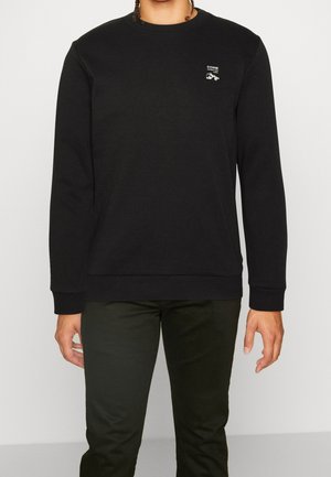 Persona que lleva una sudadera negra de manga larga con cuello redondo, con un pequeño texto blanco y un gráfico en el lado izquierdo del pecho, combinada con pantalones oscuros.
