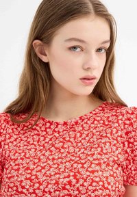 Top de patrón floral rojo con mangas cortas abullonadas, que presenta detalles en blanco. La tela parece texturizada y tiene un escote redondo.