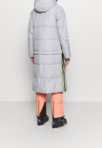 Personne portant un manteau long matelassé gris clair avec des accents jaune néon et noir, un pantalon rose saumon et des bottes de randonnée noires, vue de dos.