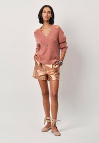 Lichtroze v-hals trui met blootgestelde schouders; glanzende roségouden shorts; beige flats met strikjes; minimalistische sieraden.