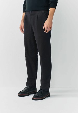 Pantalon noir à jambes droites en tissu lisse, avec fermeture à bouton et taille élastique, associé à des chaussures noires à lacets.