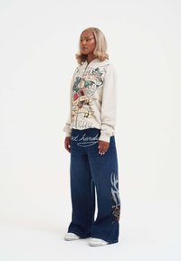 Huppari vaaleassa beigessä, jossa on graafinen printti sydämistä ja bannerista. Siniset leveälahkeiset farkut, joissa on monimutkaista brodeerausta ja valkoiset tennarit.