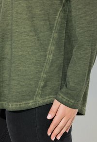 Olijfgroene langmouwtop van zacht materiaal, met een ronde zoom, zijnaden en een relaxte pasvorm. Gepaard met zwarte denim.