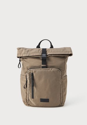 Sac à dos à rabat en tissu kaki avec une poche zippée à l'avant, des accessoires noirs et une poignée supérieure pour un transport facile. Texture lisse.