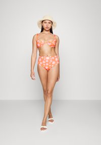 Conjunto de bikini floral laranja com detalhe em laço no top e parte de baixo de cintura alta, combinado com um chapéu de palha trançado e sandálias brancas.