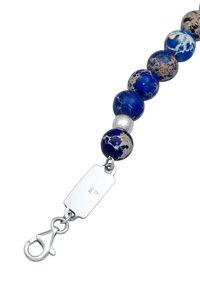 Bracelet en perles avec des pierres bleues et mouchetées, fermoir en argent et étiquette. Perles circulaires avec des textures et des motifs variés.