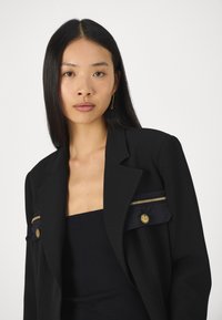 Blazer noir en tissu structuré, doté d'accents en matériel doré, de poches zippées et d'une silhouette cintrée.