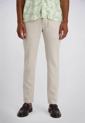Man die beige slim-fit broek, donkerbruine leren loafers en een groen geruite blouse draagt, staand tegen een effen achtergrond.