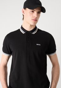 Schwarzes Polohemd aus strukturiertem Stoff mit einer drei-Knopf-Blende und einem hell gestreiften Kragen. Kleines "BOSS"-Logo auf der Brust.