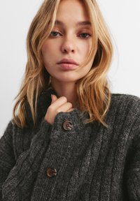 Cardigan grigio lavorato a maglia con colletto alto, caratterizzato da una texture a coste e due bottoni rotondi marroni. La mano del modello è vicino al colletto.