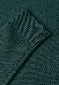 Tissu tricoté vert foncé avec une manche longue montrant un détail de poignet côtelé sur une surface plane.