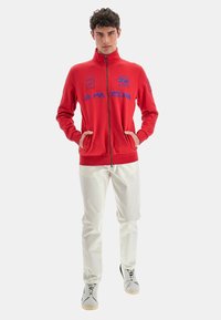 Giacca rossa con zip e ricami blu, caratterizzata da un colletto alto, due tasche laterali e orlo a coste. Abbinata a pantaloni bianchi e sneakers.